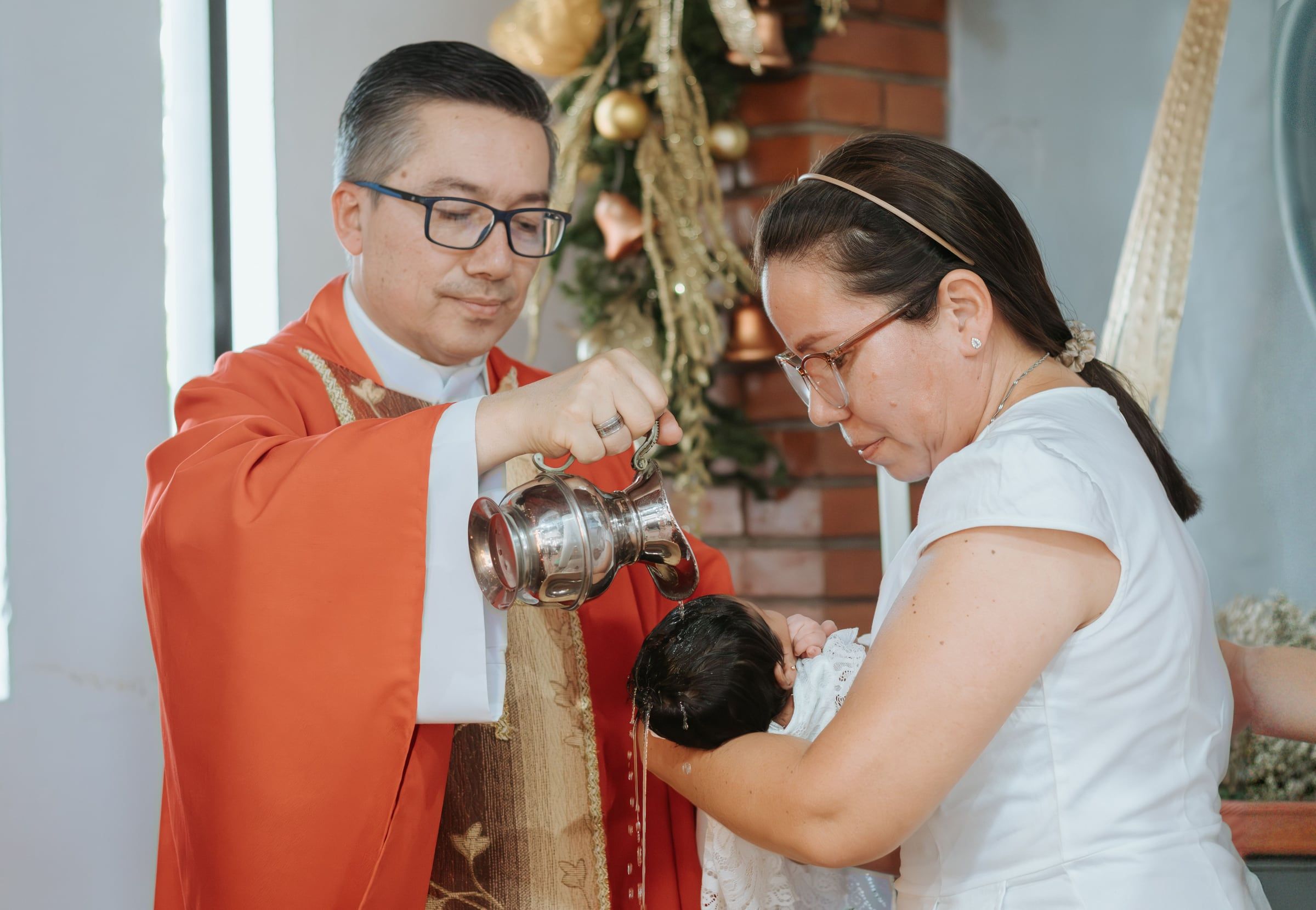 O padre de vestes vermelhas deita água sobre o bebé, ao colo da mãe.