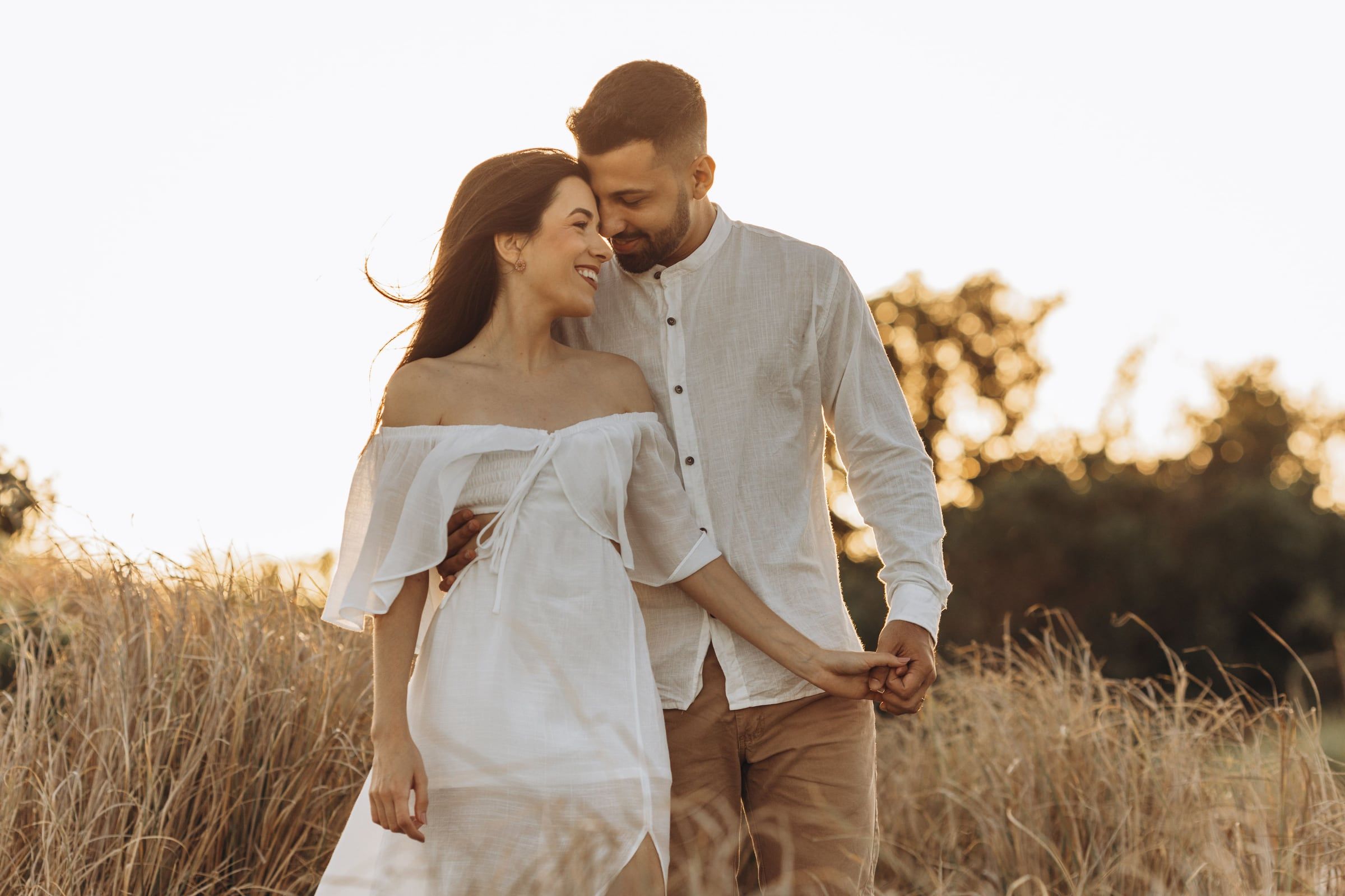 Casal num campo de erva seca ao fim da tarde, ela de vestido branco, a sorrirem um para o outro.