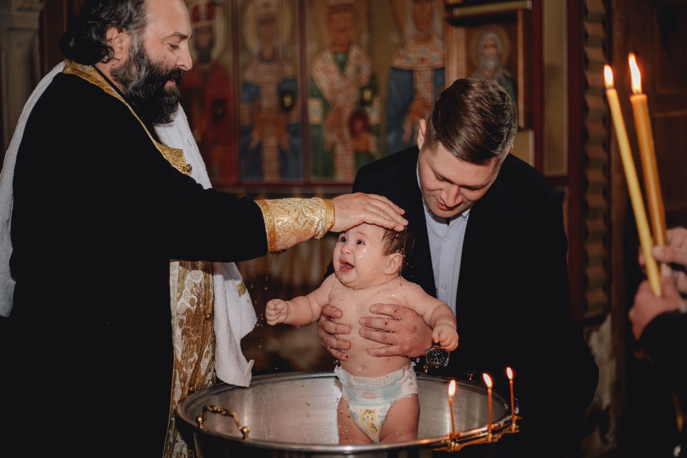 O padre segura o bebé sobre a pia batismal; o pai ao lado, velas acesas ao fundo.