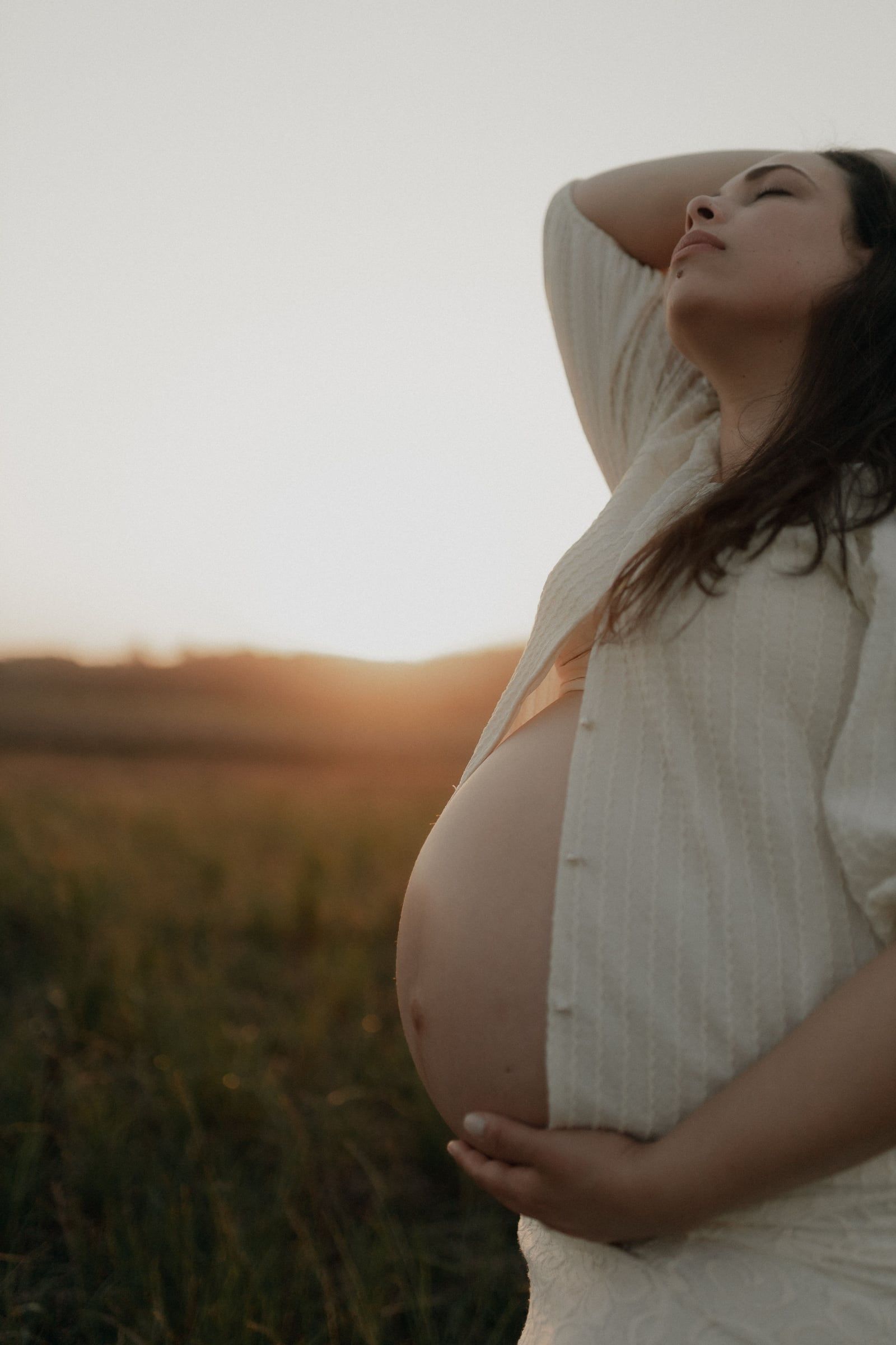 Mulher grávida de camisa aberta, de olhos fechados, num campo ao fim da tarde.